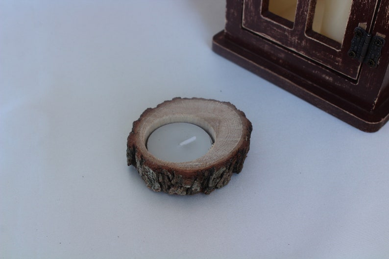 May include: A wooden candle holder made from a tree slice with a natural bark edge. The holder has a small, round indentation for a tea light candle.