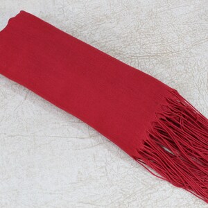 May include: A solid red scarf with a long fringe. The scarf is folded in half and laid flat on a textured surface.