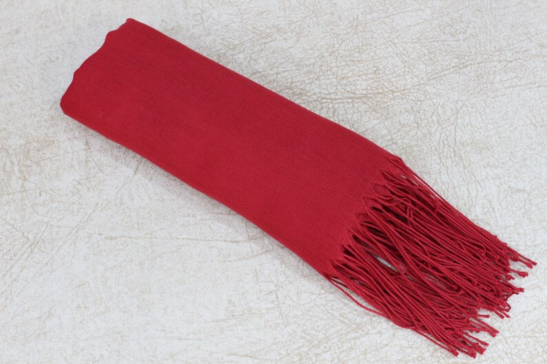 May include: A solid red scarf with a long fringe. The scarf is folded in half and laid flat on a textured surface.