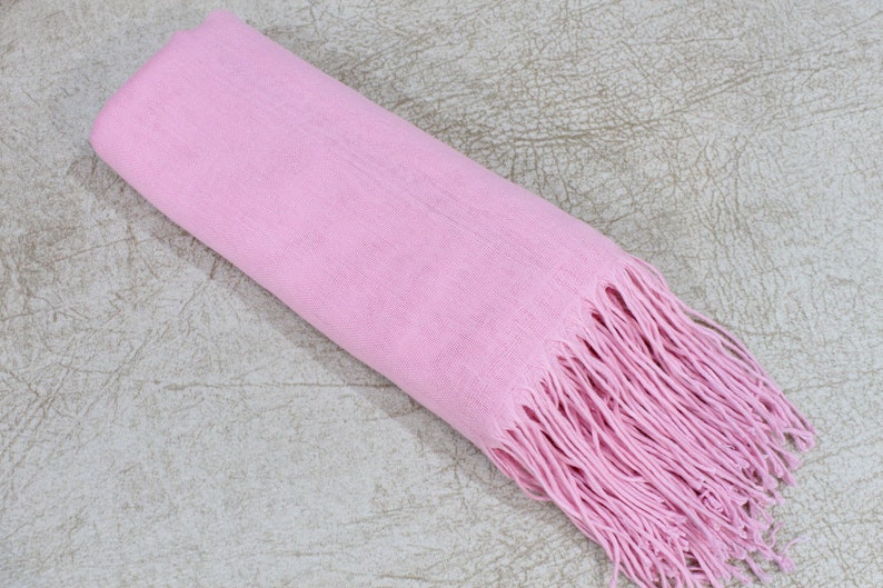 May include: A soft pink scarf with long fringe on the end. The scarf is rolled up and lying on a textured surface.