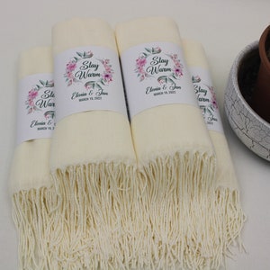 May include: Four ivory white pashmina scarves with fringed edges. Each scarf has a white label with a floral design and the text "Stay Warm" and "Elevia & Jan" with a date.