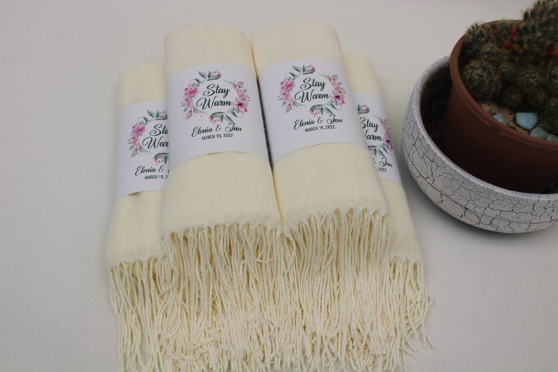 May include: Four ivory white pashmina scarves with fringed edges. Each scarf has a white label with a floral design and the text "Stay Warm" and "Elevia & Jan" with a date.