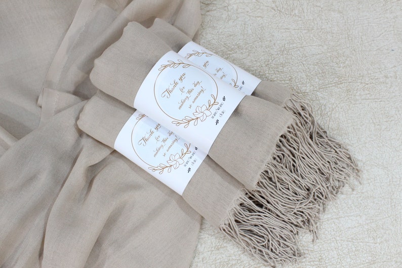 K&ouml;nnte beinhalten: Zwei beige Pashmina-Schals mit Fransen, umwickelt mit wei&szlig;en Geschenkanh&auml;ngern, auf denen in goldener Schrift "Vielen Dank, dass Sie diesen Tag so wunderbar gemacht haben!" steht.