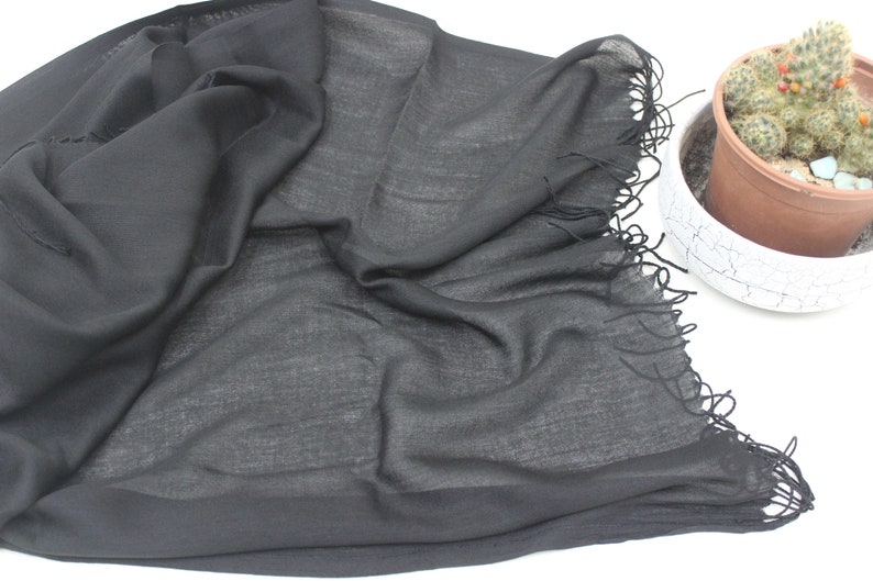 May include: A black sheer scarf with a fringed edge. The scarf is folded and laid out on a white surface.