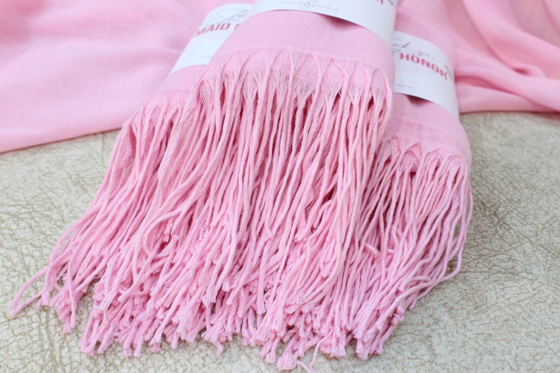 May include: Two pink pashmina scarves with fringe, folded together. The scarves have tags that say "Maid of Honor" and "Bride".