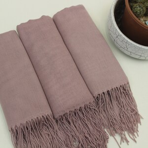 May include: Three mauve-colored scarves with fringe edges. The scarves are folded and arranged on a light beige surface.
