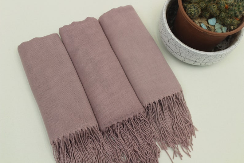 May include: Three mauve-colored scarves with fringe edges. The scarves are folded and arranged on a light beige surface.