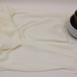 May include: A white pashmina shawl with fringed edges. The shawl is folded and laid flat on a white surface.