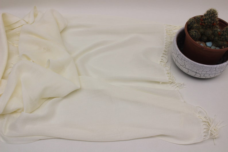 May include: A white pashmina shawl with fringed edges. The shawl is folded and laid flat on a white surface.