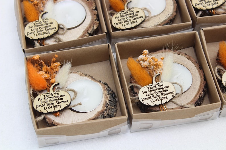 K&ouml;nnte beinhalten: Babyparty-Gastgeschenke mit einem Kerzenhalter aus Holzscheiben und einer wei&szlig;en Kerze. Jedes Gastgeschenk enth&auml;lt Trockenblumen, ein K&uuml;rbis-Etikett aus Holz mit Text und ist in einer kleinen Schachtel verpackt.