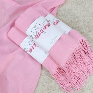 May include: Two pink pashmina scarves with fringe, wrapped together with a white tag that says "Thank you for being my Maid of Honor".