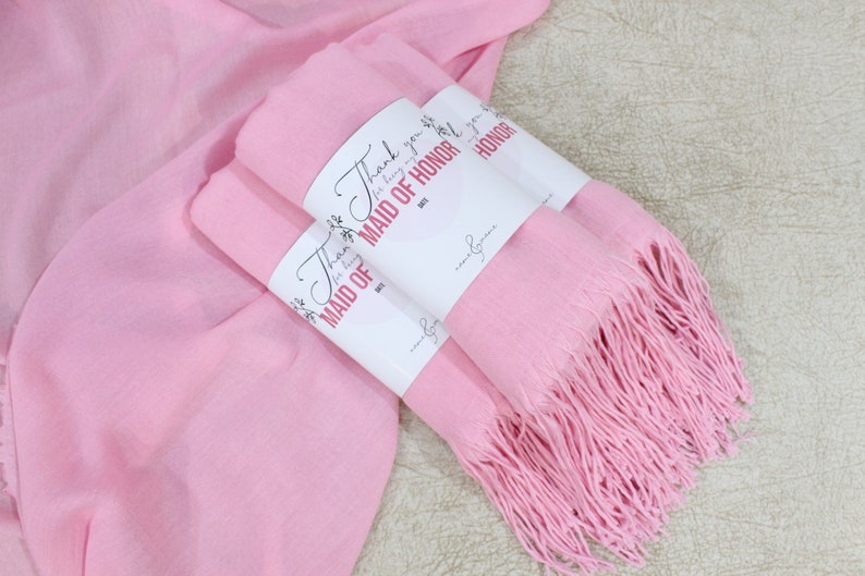 May include: Two pink pashmina scarves with fringe, wrapped together with a white tag that says "Thank you for being my Maid of Honor".