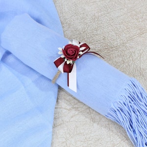 Pu&ograve; includere: Una sciarpa di pashmina blu chiaro con un accento di fiore bordeaux e bianco. La sciarpa &egrave; arrotolata e legata con un nastro.