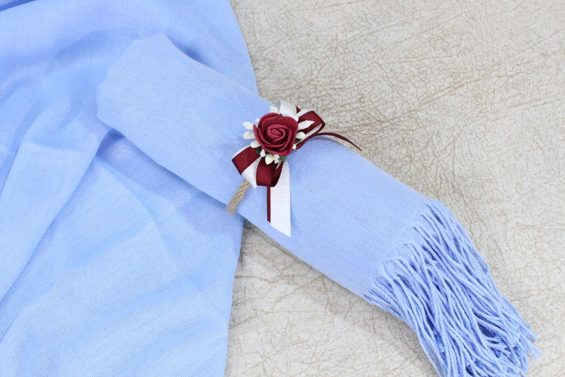 Pu&ograve; includere: Una sciarpa di pashmina blu chiaro con un accento di fiore bordeaux e bianco. La sciarpa &egrave; arrotolata e legata con un nastro.