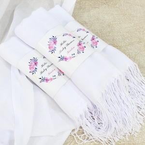 May include: Three white scarves with fringed edges, each wrapped with a floral design paper band that says "Hello Baby Shower" and a name.
