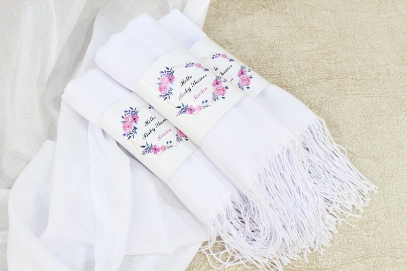 May include: Three white scarves with fringed edges, each wrapped with a floral design paper band that says "Hello Baby Shower" and a name.