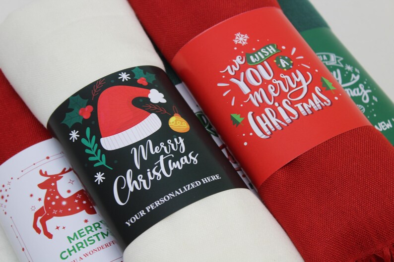 Pode incluir: Tr&ecirc;s argolas de guardanapo com tema natalino. Uma &eacute; vermelha com texto branco que diz "We wish you a merry Christmas". Outra &eacute; preta com texto branco que diz "Merry Christmas" e um chap&eacute;u de Papai Noel vermelho e branco. A &uacute;ltima &eacute; branca com texto vermelho e verde que diz "Merry Christmas" e uma rena vermelha.