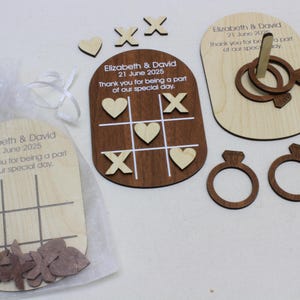 K&ouml;nnte beinhalten: Holz-Hochzeitsgeschenke mit Tic-Tac-Toe-Spielen, Ringwurfspielen und ringf&ouml;rmigen Ausschnitten. Die Gastgeschenke enthalten gravierten Text mit einem Datum und einer Dankesbotschaft. Die Artikel befinden sich in einem transparenten wei&szlig;en Beutel.