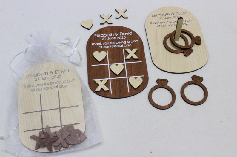 K&ouml;nnte beinhalten: Holz-Hochzeitsgeschenke mit Tic-Tac-Toe-Spielen, Ringwurfspielen und ringf&ouml;rmigen Ausschnitten. Die Gastgeschenke enthalten gravierten Text mit einem Datum und einer Dankesbotschaft. Die Artikel befinden sich in einem transparenten wei&szlig;en Beutel.