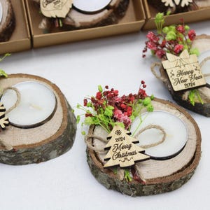 May include: Three wooden candle holders with small tea lights, decorated with sprigs of red and green flowers and a wooden snowflake or Christmas tree ornament. The ornaments have the text "Happy New Year 2024" or "2024 Merry Christmas".
