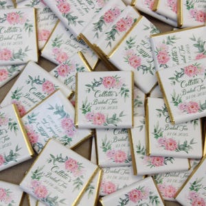 Puede incluir: Un mont&oacute;n de peque&ntilde;as barras de chocolate blanco con envoltorios de papel de aluminio dorado. Cada barra tiene un dise&ntilde;o floral con flores rosas y hojas verdes. El texto "Collette's Bridal Tea 31.06.2025" est&aacute; impreso en cada barra.