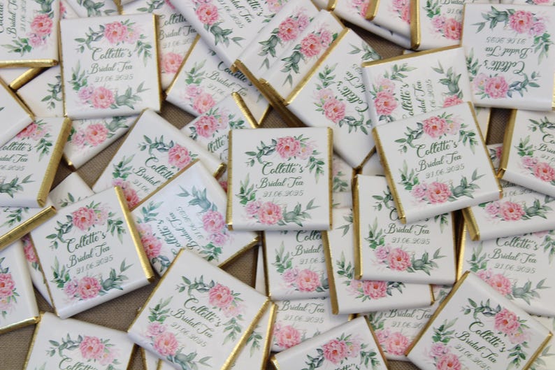 Puede incluir: Un mont&oacute;n de peque&ntilde;as barras de chocolate blanco con envoltorios de papel de aluminio dorado. Cada barra tiene un dise&ntilde;o floral con flores rosas y hojas verdes. El texto "Collette's Bridal Tea 31.06.2025" est&aacute; impreso en cada barra.