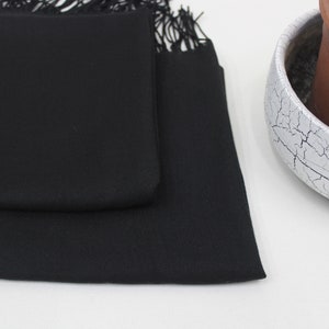 May include: A black pashmina scarf with fringe. The scarf is folded in half and is lying on a white surface.