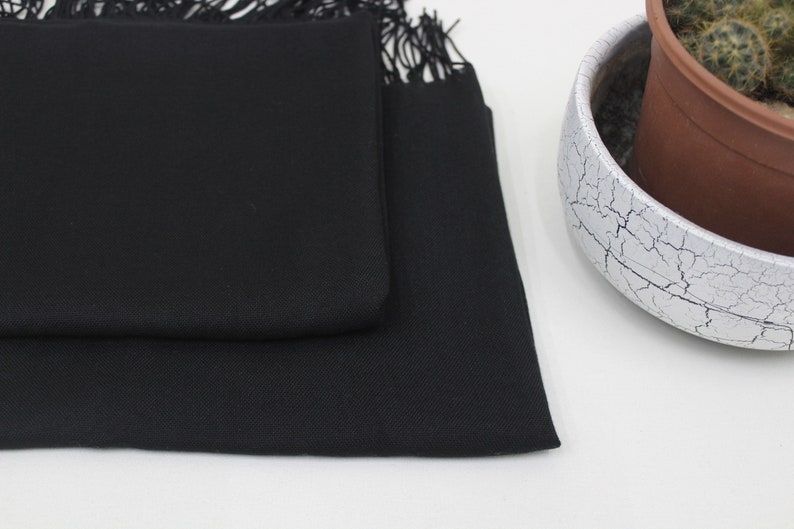 May include: A black pashmina scarf with fringe. The scarf is folded in half and is lying on a white surface.