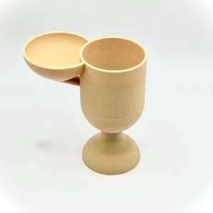 May include: A beige, cup-shaped object with a small, attached bowl. The cup has a rounded base and a cylindrical top. The bowl extends from the side of the cup. The object is made of a textured material and sits on a white surface.