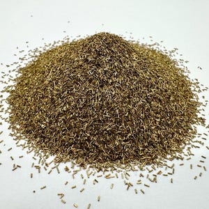 May include: A pile of small, cylindrical, brass-colored metal pieces. The pieces are scattered on a white surface, with the main concentration forming a small mound. The overall appearance is of a collection of small, uniform components.