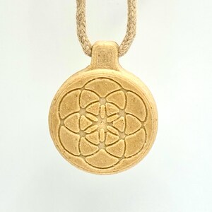May include: A beige pendant necklace with a circular, engraved design. The pendant features a floral pattern and hangs from a beige rope. The necklace is centered against a white background.