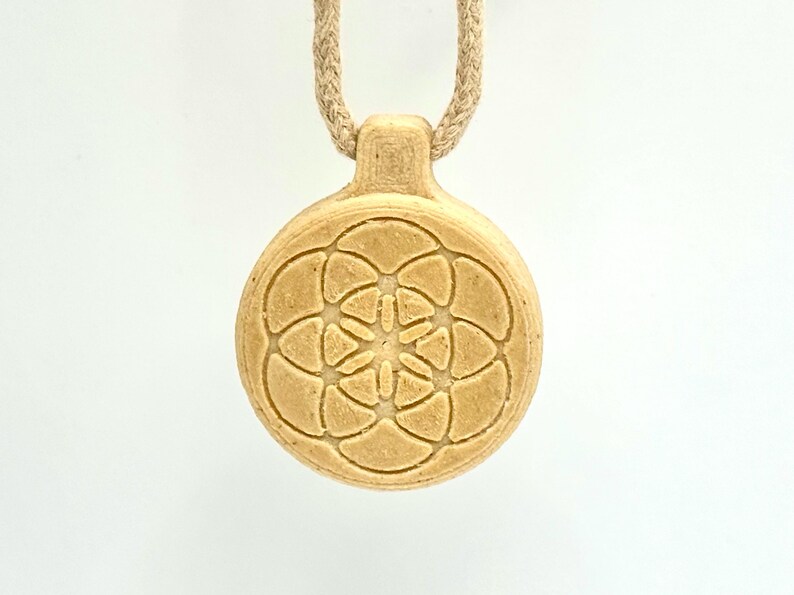 May include: A beige pendant necklace with a circular, engraved design. The pendant features a floral pattern and hangs from a beige rope. The necklace is centered against a white background.