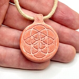 Op de afbeelding: Een ronde, koraalkleurige hanger met een geometrisch bloemmotief, hangend aan een beige touw. De hanger wordt in een hand gehouden tegen een witte achtergrond. Het ontwerp van de hanger heeft een symmetrisch patroon.