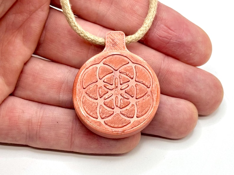 Op de afbeelding: Een ronde, koraalkleurige hanger met een geometrisch bloemmotief, hangend aan een beige touw. De hanger wordt in een hand gehouden tegen een witte achtergrond. Het ontwerp van de hanger heeft een symmetrisch patroon.