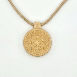 May include: A tan pendant necklace with a circular pendant displaying a flower of life pattern. The pendant hangs from a braided, tan-coloured cord. The necklace is set against a white background.