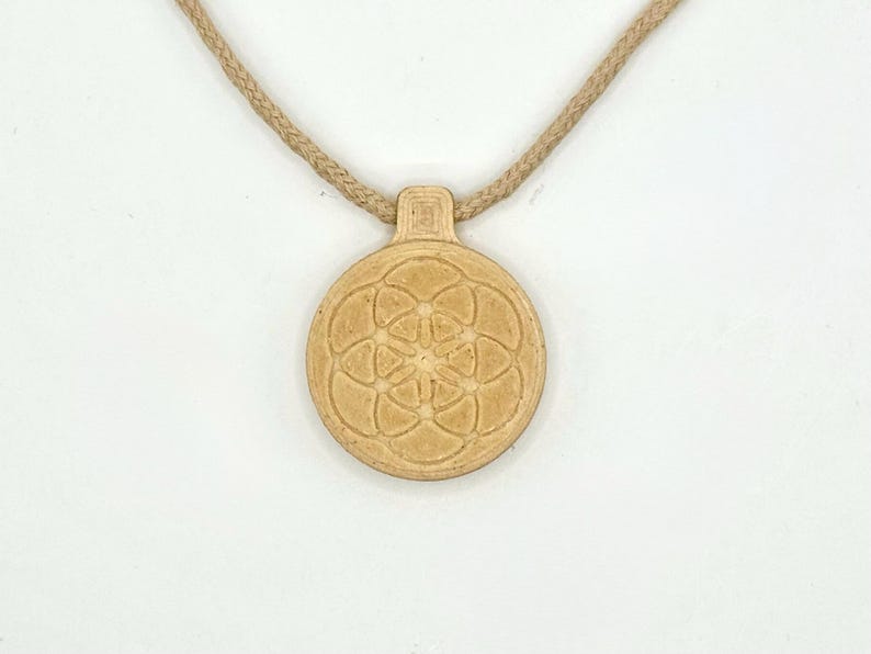 May include: A tan pendant necklace with a circular pendant displaying a flower of life pattern. The pendant hangs from a braided, tan-coloured cord. The necklace is set against a white background.