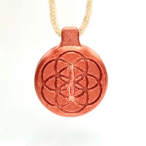 Op de afbeelding: Een roodbruine hangerketting met een cirkelvormig, gegraveerd ontwerp met een geometrisch bloemmotief. De hanger is bevestigd aan een beige koord. De ketting wordt gepresenteerd tegen een witte achtergrond.