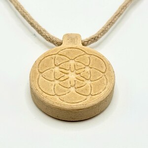 May include: A beige pendant necklace featuring a circular, floral design. The pendant is attached to a beige, rope-style cord. The pendant has a raised, textured pattern and a small rectangular top.