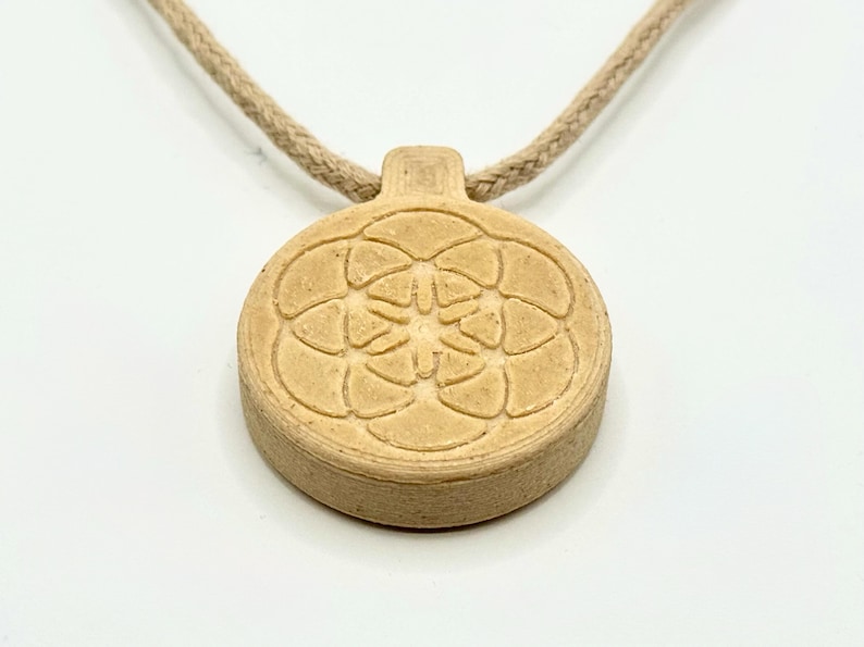 May include: A beige pendant necklace featuring a circular, floral design. The pendant is attached to a beige, rope-style cord. The pendant has a raised, textured pattern and a small rectangular top.