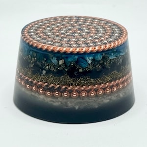 May include: A cylindrical orgone energy generator with a copper-colored top featuring a geometric pattern. The sides are layered with blue and black crystals, copper wire, and a copper-colored beaded band. The base is a gradient of gray and dark blue.