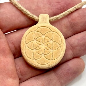 May include: A beige pendant necklace with a circular pendant displaying a floral pattern. The pendant hangs from a beige cord. The necklace is held in a person's hand.