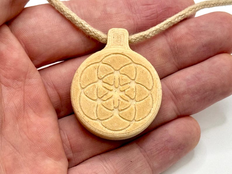 May include: A beige pendant necklace with a circular pendant displaying a floral pattern. The pendant hangs from a beige cord. The necklace is held in a person's hand.