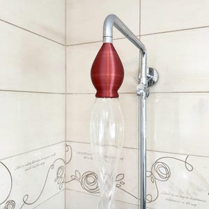 Könnte beinhalten: Einzigartiger Duschkopf mit einem klaren, länglichen Glaskörper und einer bordeauxfarbenen Oberseite. Der Duschkopf ist an einem verchromten Duscharm befestigt und an einer gefliesten Wand montiert. Der Glaskörper hat ein gedrehtes Design.