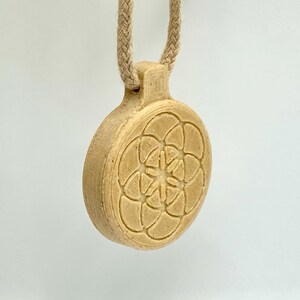 May include: A beige pendant with a floral pattern, hanging from a braided cord. The pendant is circular and features a raised, symmetrical design. The cord is attached to the pendant via a small loop at the top.
