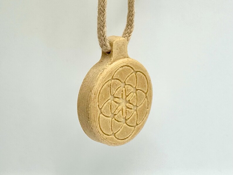 May include: A beige pendant with a floral pattern, hanging from a braided cord. The pendant is circular and features a raised, symmetrical design. The cord is attached to the pendant via a small loop at the top.