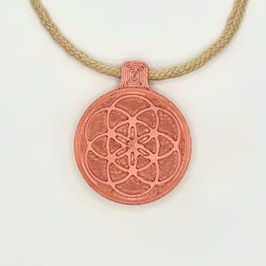 Op de afbeelding: Een koperkleurige hangerketting met een cirkelvormige hanger met een gedetailleerd bloemmotief. De hanger hangt aan een beige, touwachtig koord. De ketting wordt gepresenteerd tegen een witte achtergrond.