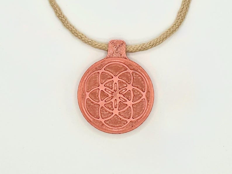 Op de afbeelding: Een koperkleurige hangerketting met een cirkelvormige hanger met een gedetailleerd bloemmotief. De hanger hangt aan een beige, touwachtig koord. De ketting wordt gepresenteerd tegen een witte achtergrond.