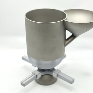 Könnte beinhalten: Ein zylindrischer Behälter aus mattem silberfarbenem Metall mit einem seitlich angebrachten Trichter, montiert auf einem vierzackigen Sockel. Die Struktur weist eine glatte, gleichmäßige Oberfläche und eine vertikale Ausrichtung auf.