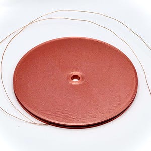 Può includere: Una bobina di nastro color rame con una superficie strutturata. Tre fili di filo sottile color rame si estendono dalla bobina. La bobina ha un foro centrale. Il nastro è di un colore marrone rossastro smorzato.