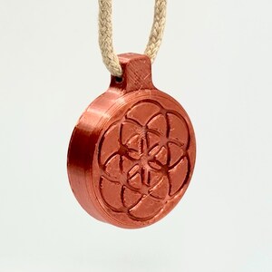 Op de afbeelding: Een koperkleurige hanger met een geometrisch bloem des levens-ontwerp, hangend aan een beige touw. De ronde hanger heeft een diameter van 3,8 cm en heeft een gedetailleerd, verhoogd patroon. Het touw is ongeveer 25 cm lang.